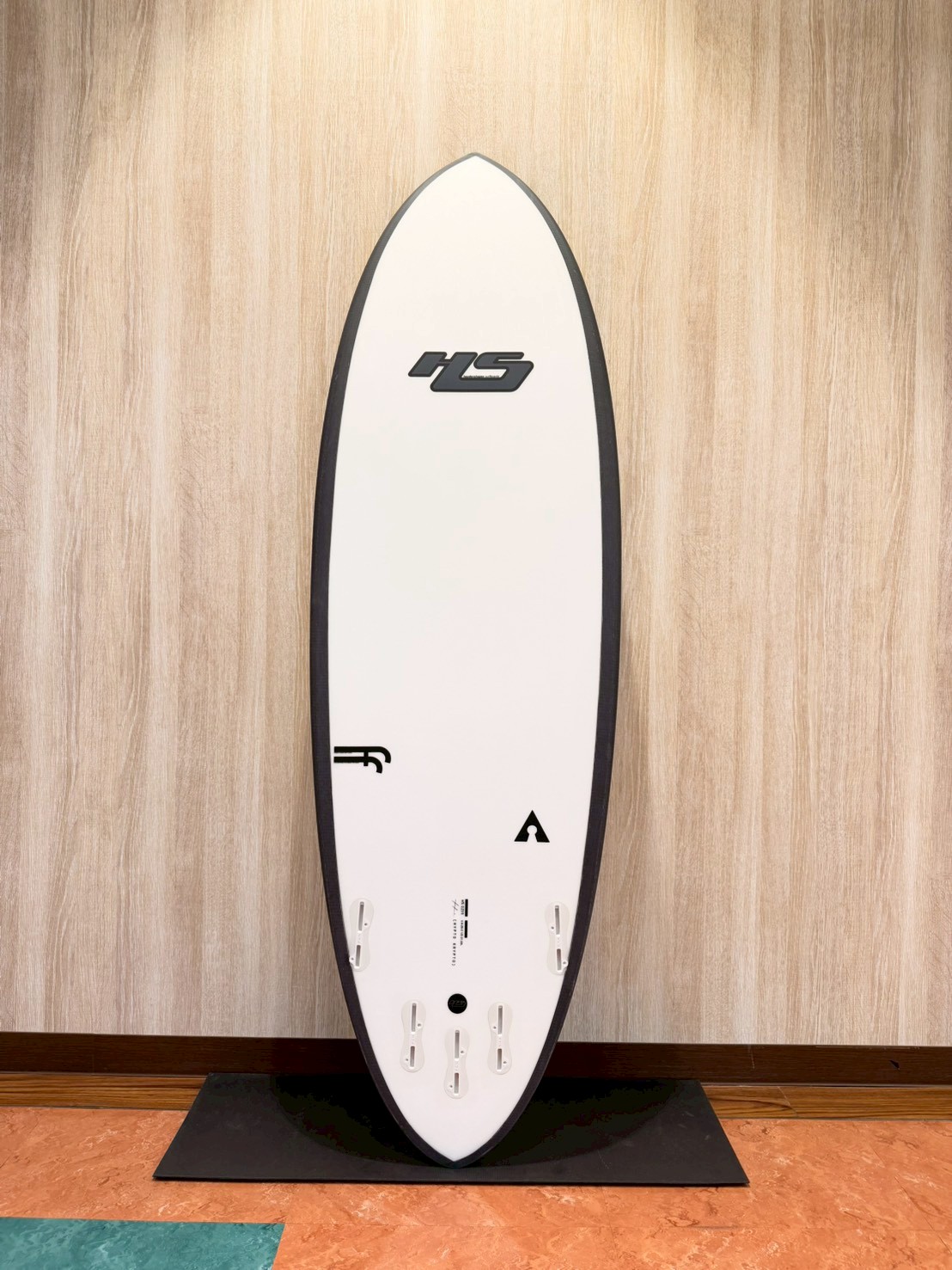 White surfboard with black rails standing upright on a black display mat against a wood-paneled wall, logo near the center and fin/foot well slots at the bottom