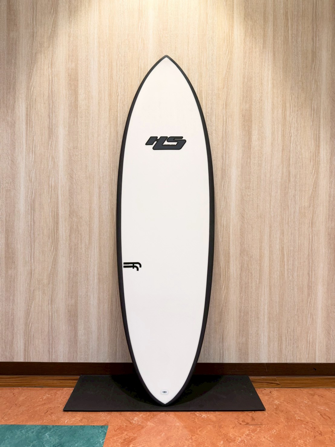 White surfboard with a black edge leaning on a black mat against a wood-paneled wall, featuring an 'HS' logo midboard.