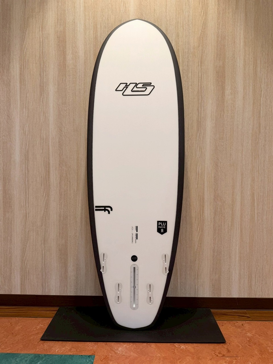 White surfboard with black edge trim, standing upright on a black mat against a wood-paneled wall; fin boxes visible near the tail.