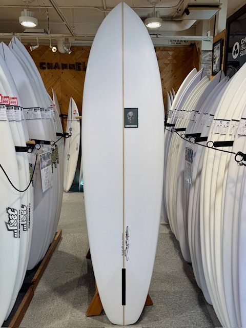 White surfboard standing upright on a wooden stand in a surf shop, surrounded by rows of other boards and a small square logo on its chest.