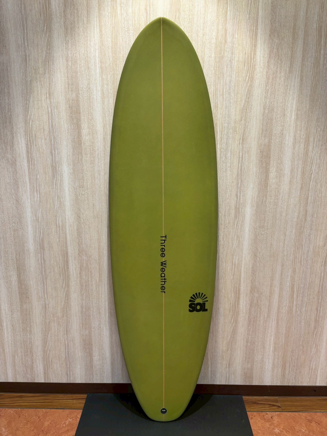 Green surfboard standing upright against a wood panel wall, with a thin center rail and SOL logo near the bottom on a black display stand.