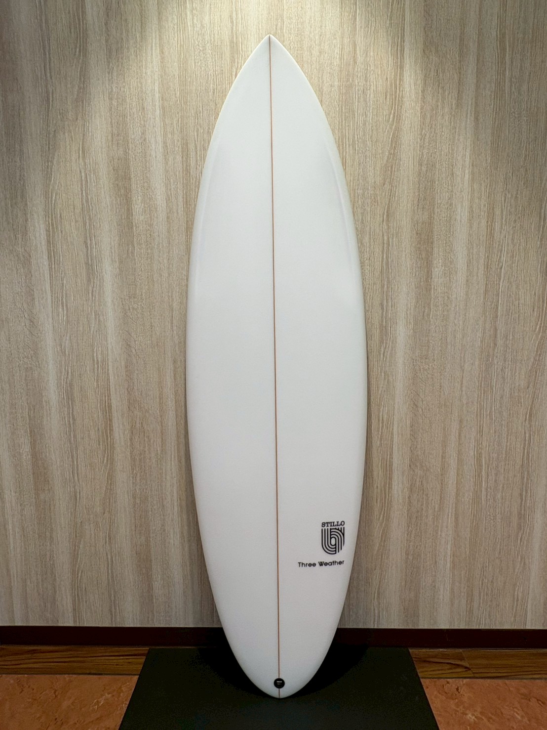 White surfboard with a thin red stringer standing upright against a wood-paneled wall on a black mat.