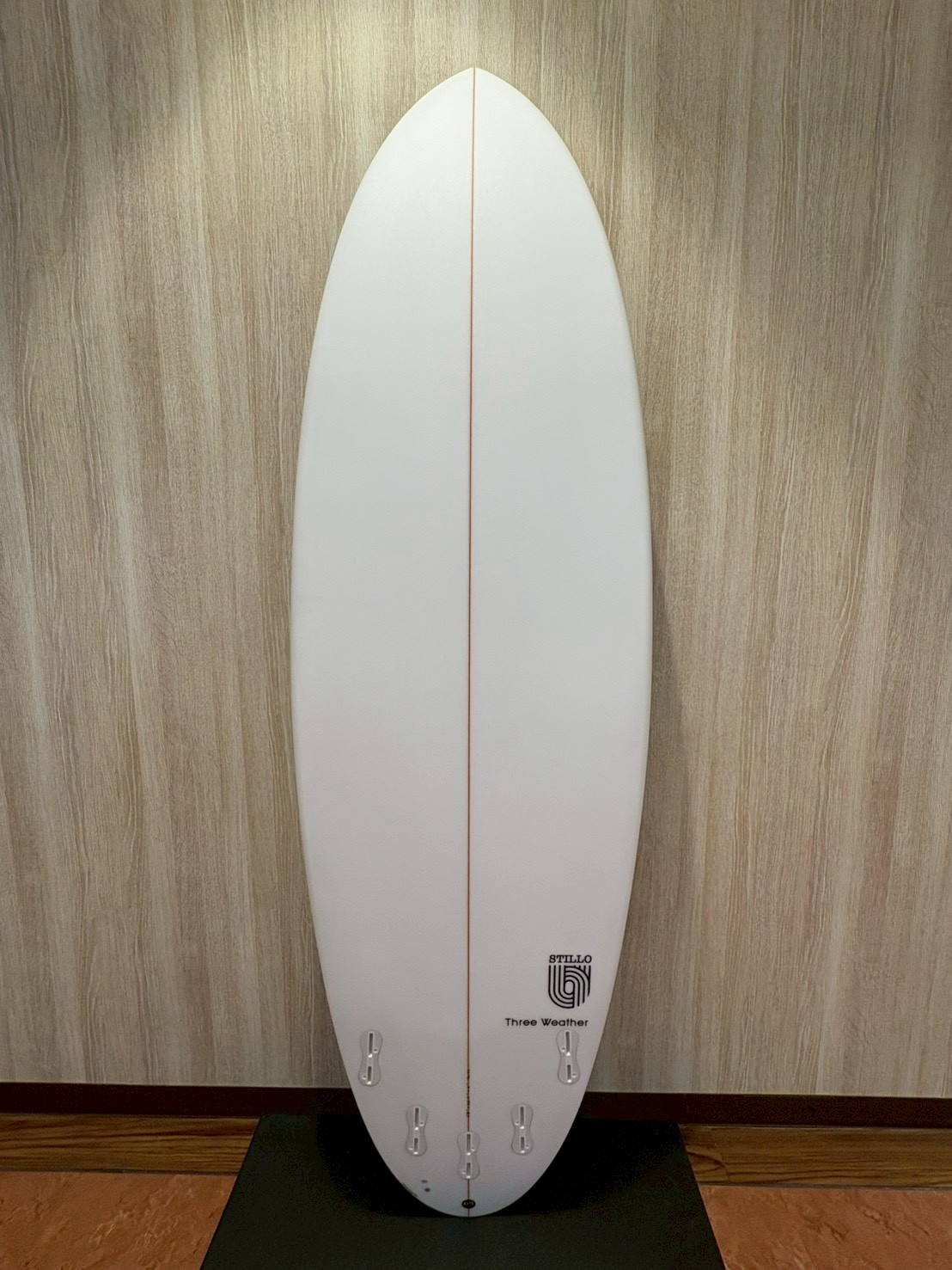 White vertical surfboard with an orange center stringer, standing on a black mat against a wood-paneled wall; tail has multiple fin boxes and a small logo near the bottom.