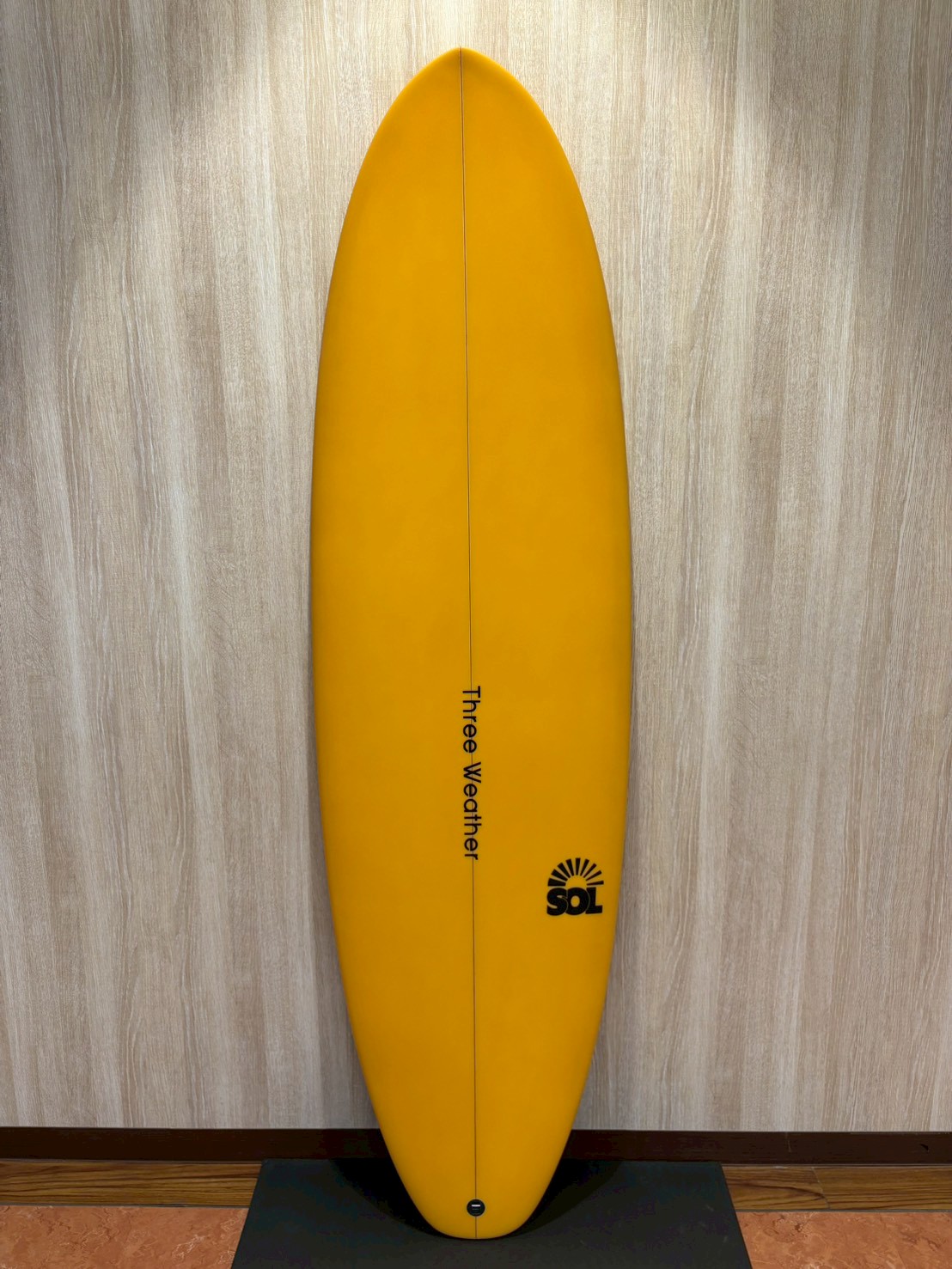 Yellow surfboard standing upright against a light wood wall, with 'Three Weather' branding along the center and a SOL logo near the bottom.