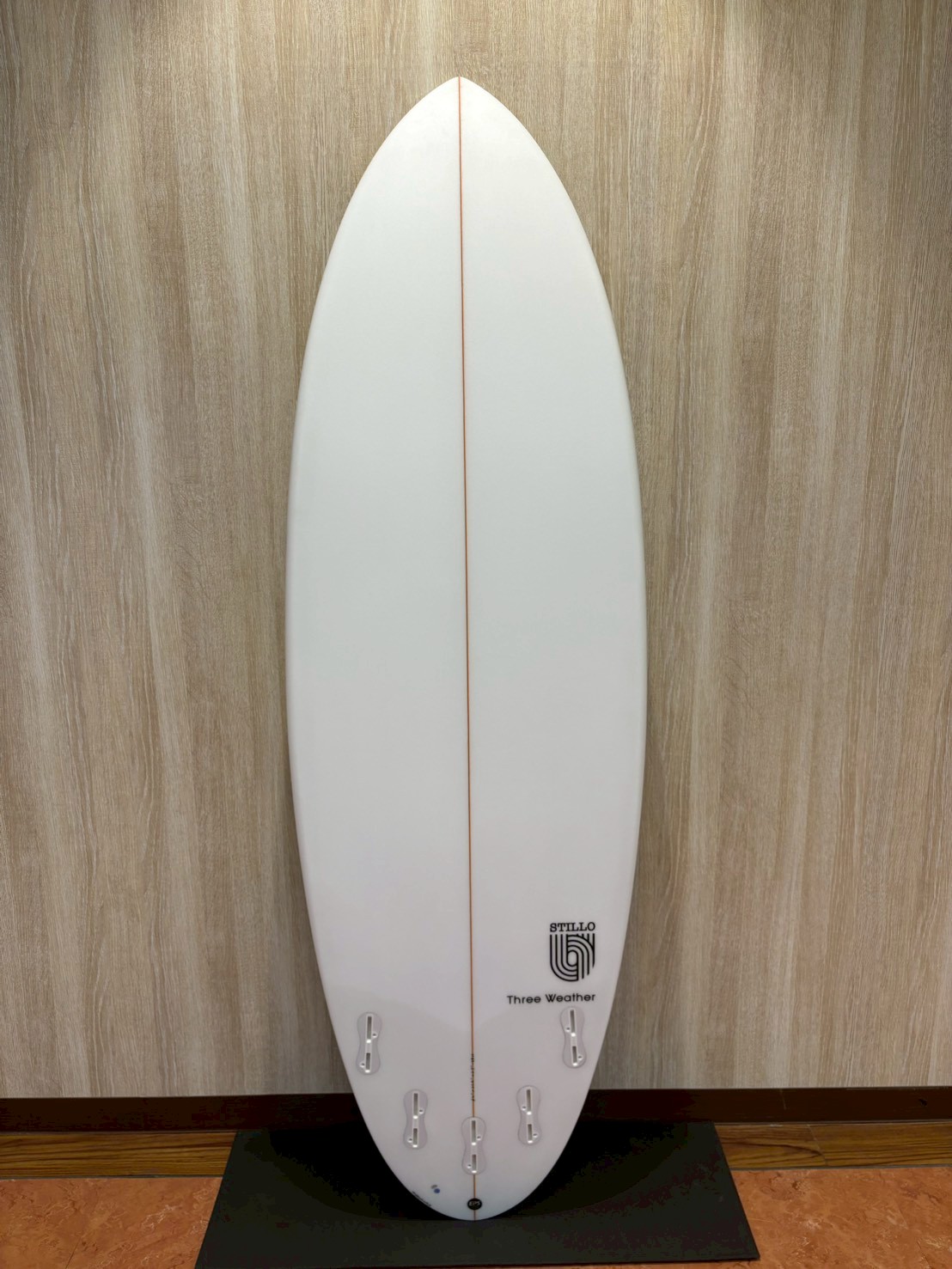 White surfboard with a thin red center stringer, standing upright against a wooden wall on a display stand.