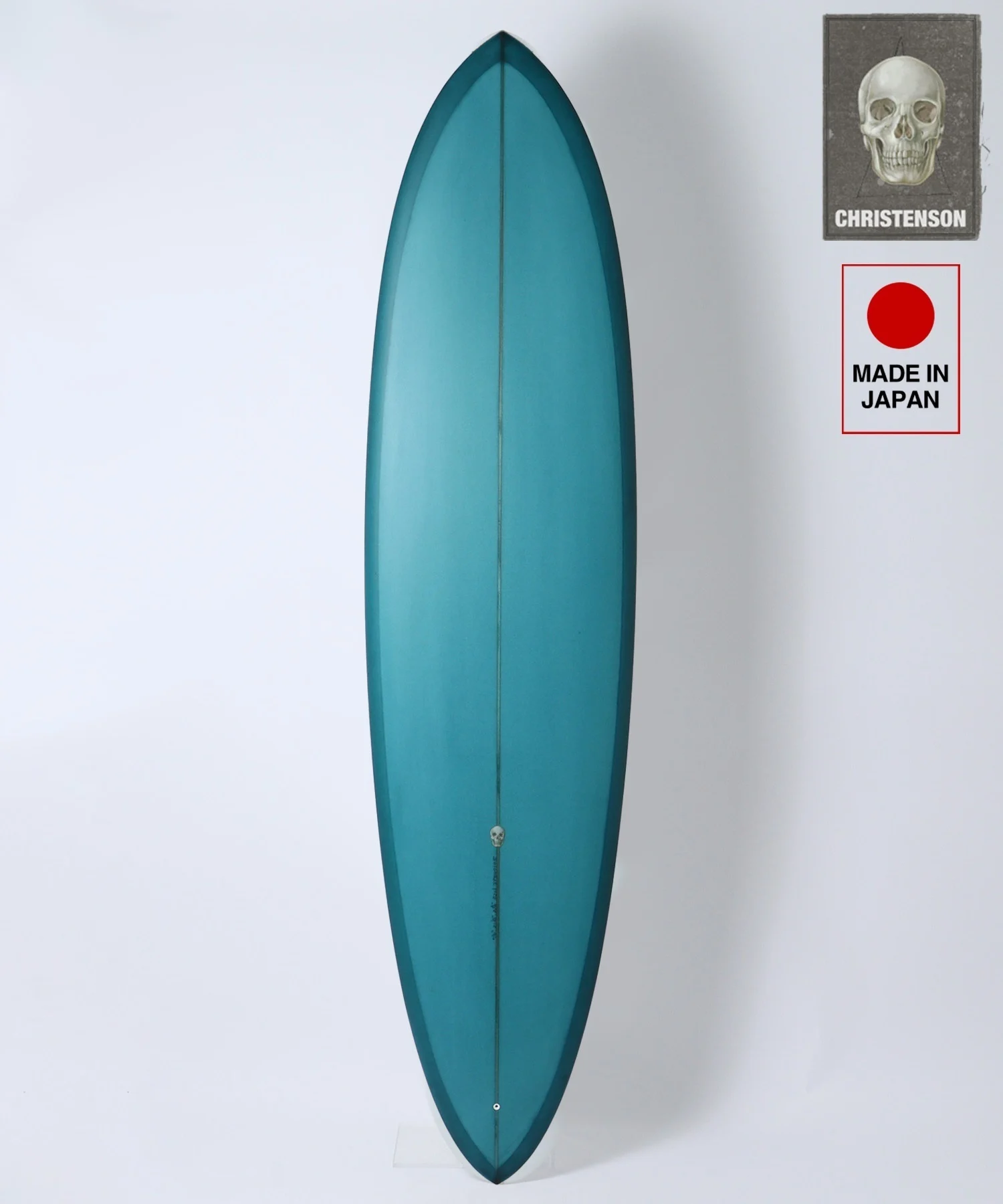 image - SHOP NEWS | ムラサキスポーツオンラインストア 通販 Vertical teal surfboard standing against a white wall, with two stickers on the upper right (skull 'Christenson' badge and 'Made in Japan' label).