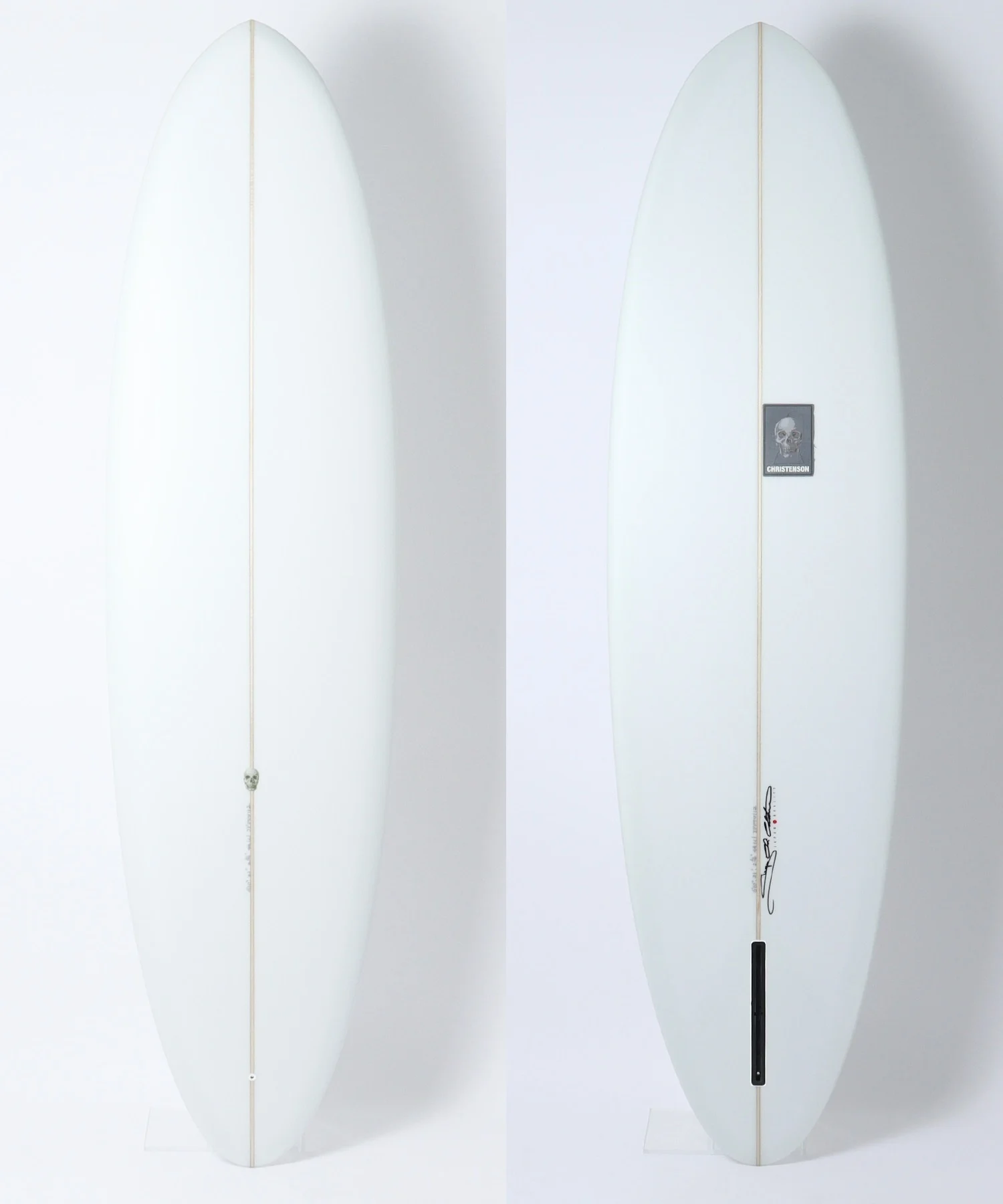 image - SHOP NEWS | ムラサキスポーツオンラインストア 通販 Two white surfboards standing upright side by side against a white backdrop; left board has a small skull charm on the cord running down its fin, right board shows a skull sticker and signature near the bottom.