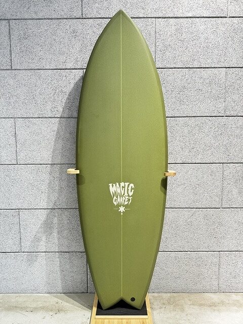 s-IMG_4245 - SHOP NEWS | ムラサキスポーツオンラインストア 通販 Olive-green surfboard standing upright on a wooden stand against a gray brick wall; white 'Magic' logo in the middle.