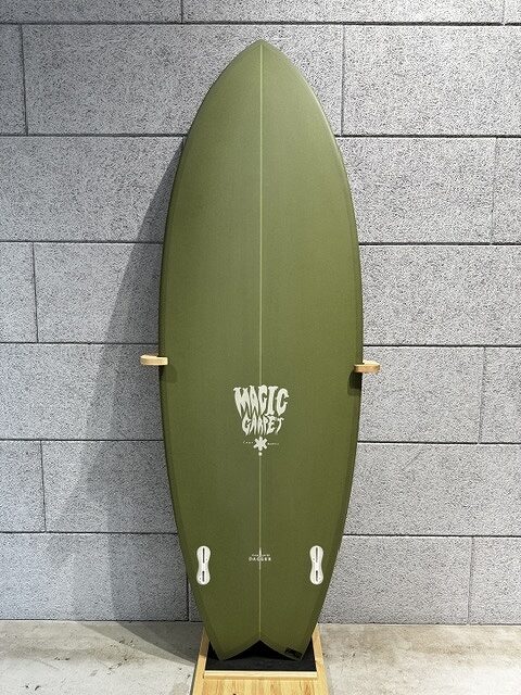 s-IMG_4246 - SHOP NEWS | ムラサキスポーツオンラインストア 通販 Olive-green surfboard with a pointed nose, standing upright on a wooden rack against a gray tiled wall, logo reads 'Magic Carvers' in the center.