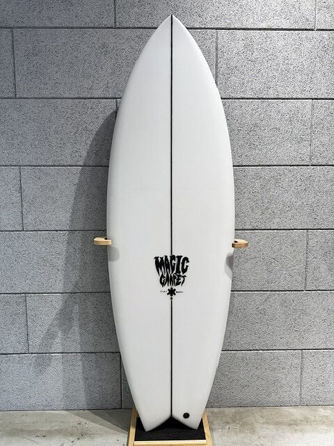 s-IMG_4247 - SHOP NEWS | ムラサキスポーツオンラインストア 通販 White surfboard with a black center seam, mounted on a wooden stand against a gray brick wall; logo near center.