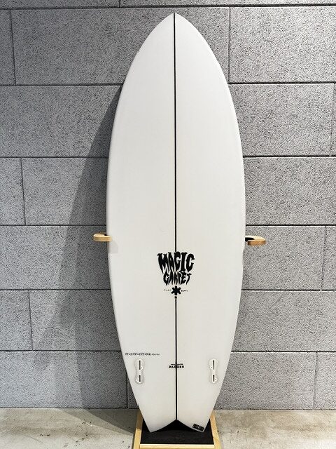 s-IMG_4248 - SHOP NEWS | ムラサキスポーツオンラインストア 通販 White surfboard with a black center stringer, resting on a wooden stand against a gray brick wall; displays the 'Magic Carpet' logo near the lower half.