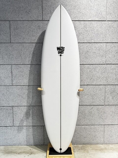 s-IMG_4249 - SHOP NEWS | ムラサキスポーツオンラインストア 通販 White surfboard standing upright against a gray brick wall on a wooden rack, featuring a black center stringer and a logo near the top.