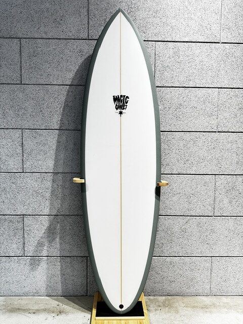 s-IMG_4251 - SHOP NEWS | ムラサキスポーツオンラインストア 通販 White surfboard with a dark green edge mounted vertically on a wooden display stand against a gray brick wall.