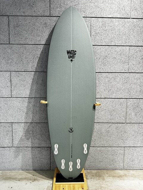 s-IMG_4252 - SHOP NEWS | ムラサキスポーツオンラインストア 通販 Gray-green surfboard standing upright on a wooden display stand against a gray brick wall.