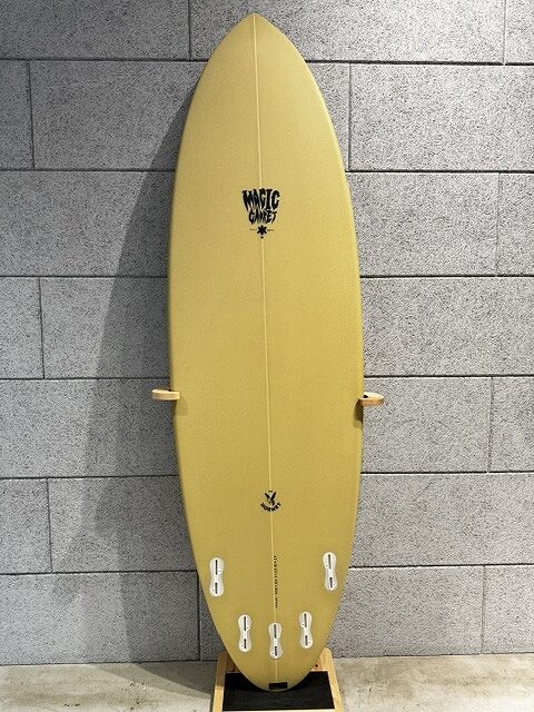 s-IMG_4254 - SHOP NEWS | ムラサキスポーツオンラインストア 通販 Yellow surfboard standing upright on a wooden rack against a gray tiled wall, with a small black logo near the top.