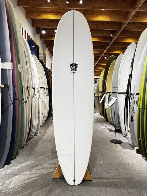 s-IMG_4255 - SHOP NEWS | ムラサキスポーツオンラインストア 通販 White surfboard standing upright on a display rack in a surf shop, with rows of colorful boards lining both sides of the aisle.
