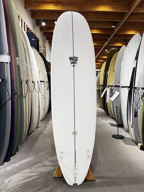 s-IMG_4256 - SHOP NEWS | ムラサキスポーツオンラインストア 通販 White surfboard standing upright on a display rack in a surf shop, surrounded by colorful boards under a wooden ceiling.