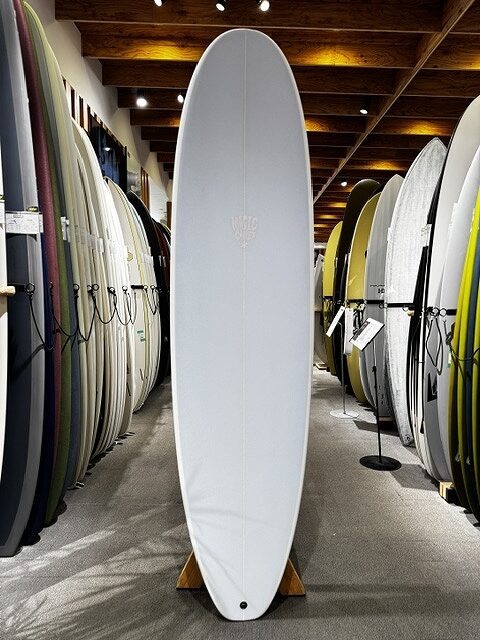 s-IMG_4257 - SHOP NEWS | ムラサキスポーツオンラインストア 通販 White surfboard standing upright on a wooden stand in a surf shop, surrounded by rows of other boards.