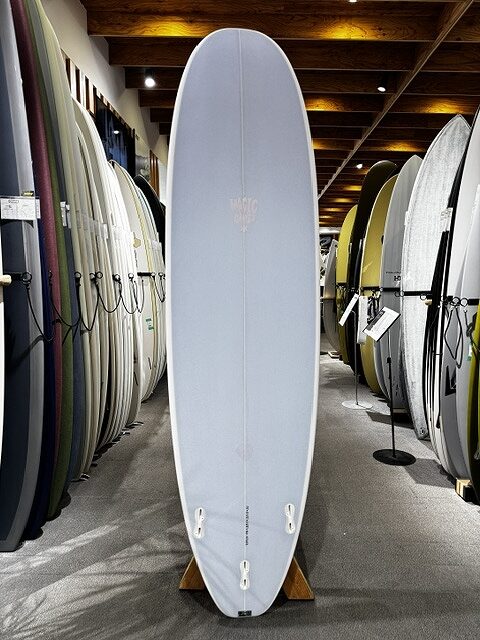 s-IMG_4258 - SHOP NEWS | ムラサキスポーツオンラインストア 通販 Light blue surfboard standing upright on a wooden stand in a surf shop, surrounded by other boards on racks.