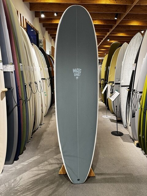 s-IMG_4259 - SHOP NEWS | ムラサキスポーツオンラインストア 通販 Gray surfboard on a wooden stand in a shop, surrounded by rows of colorful surfboards on racks.