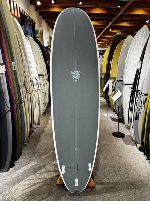s-IMG_4260 - SHOP NEWS | ムラサキスポーツオンラインストア 通販 Center-standing gray surfboard on a stand in a shop, surrounded by rows of other boards along the walls.