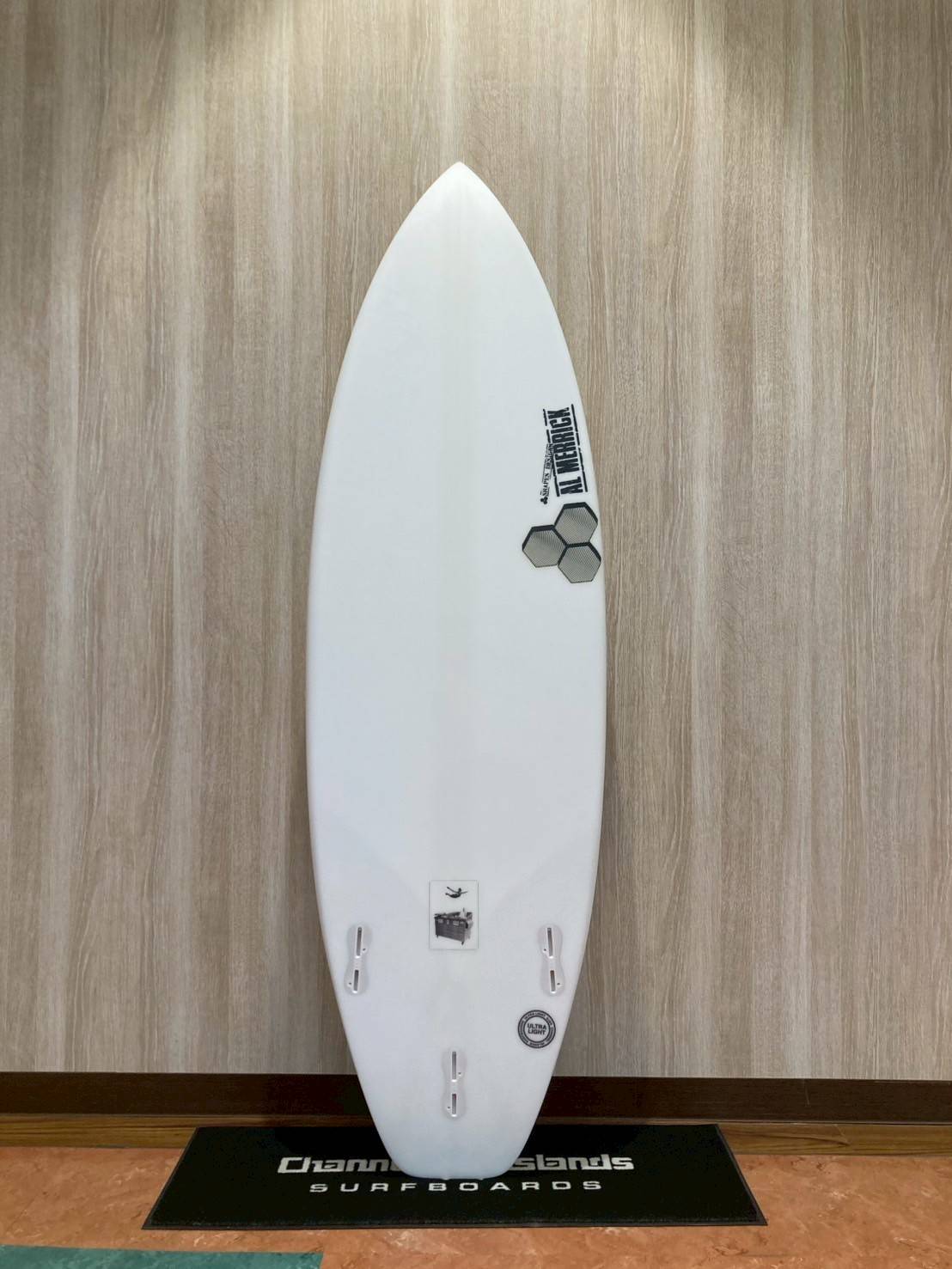 White surfboard standing upright against a wood-paneled wall on a display mat with 'Surfboards' branding.