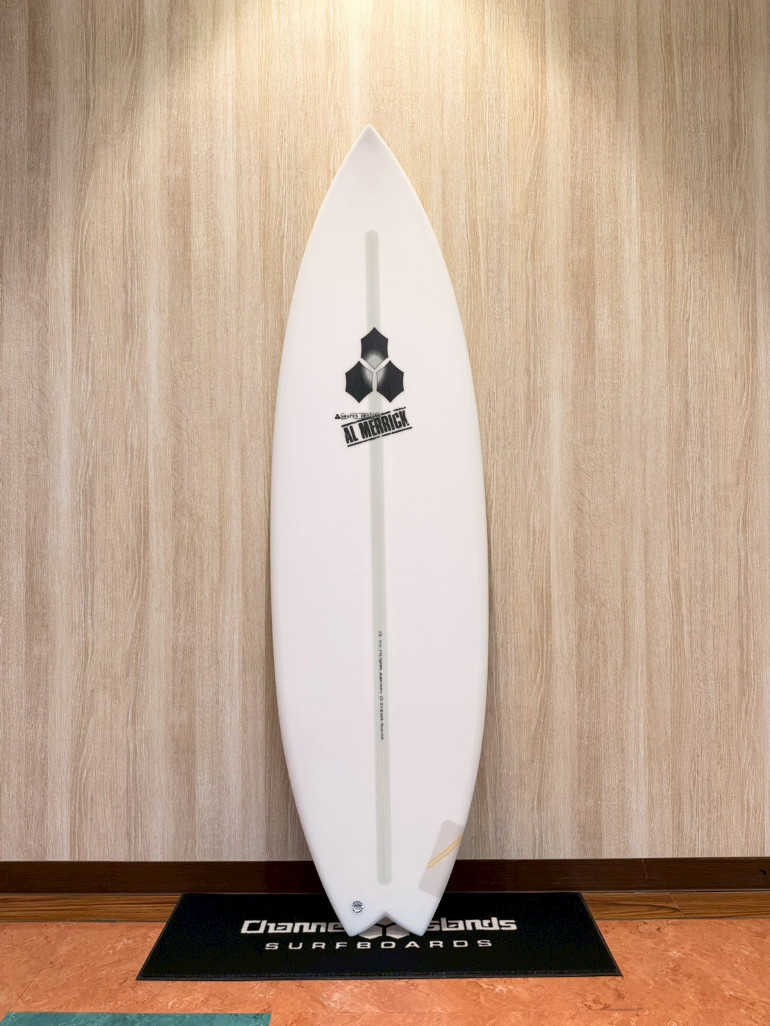 White surfboard standing upright against a wood-paneled wall with a branded black mat at its base.