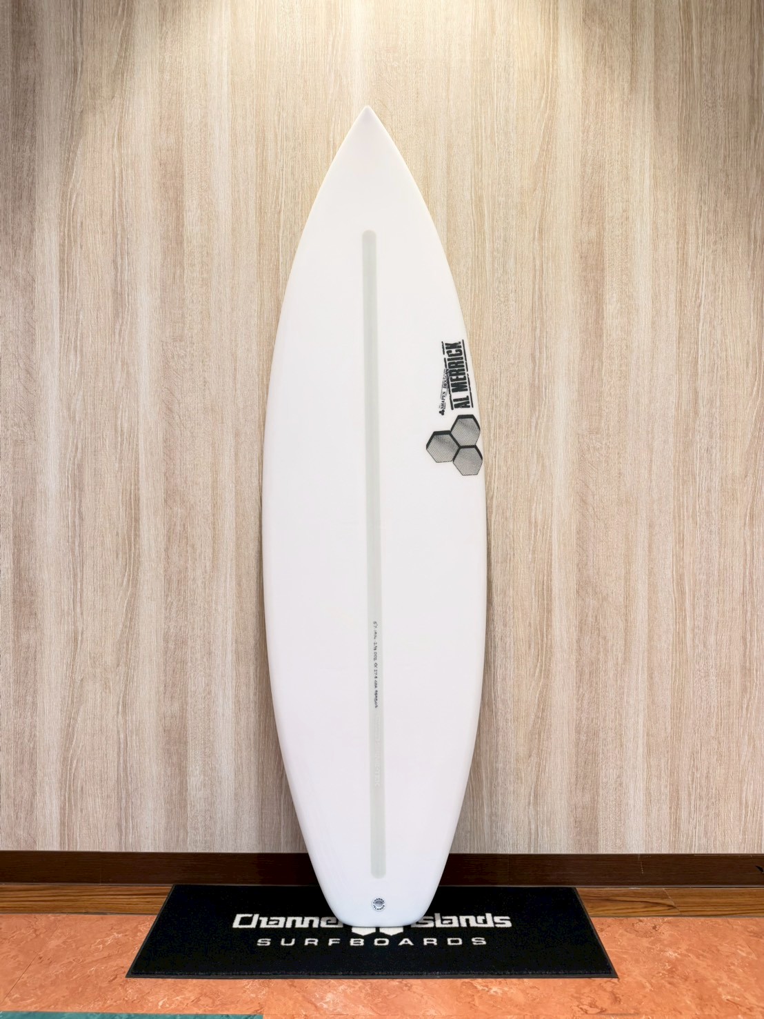 White surfboard with a pale gray stringer stands upright on a black display mat against a wood-paneled wall.
