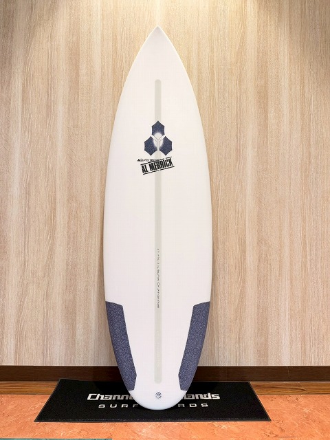 White surfboard standing upright on a black mat indoors, with blue tail pads and an Al Merrick logo center-right.