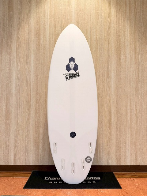 White surfboard standing upright against a light wood wall, logo near the top, four fin boxes along the bottom footer, placed on a black mat.
