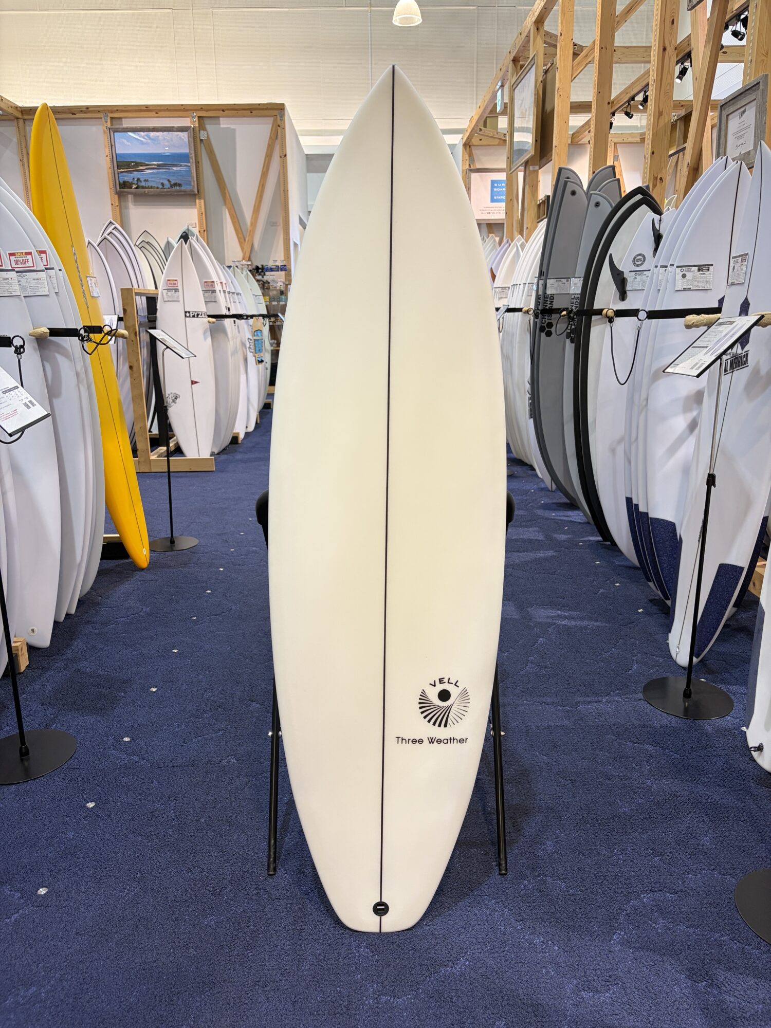 Center white surfboard with 'Three Weather' logo standing upright in a surf shop, surrounded by racks of boards on all sides.