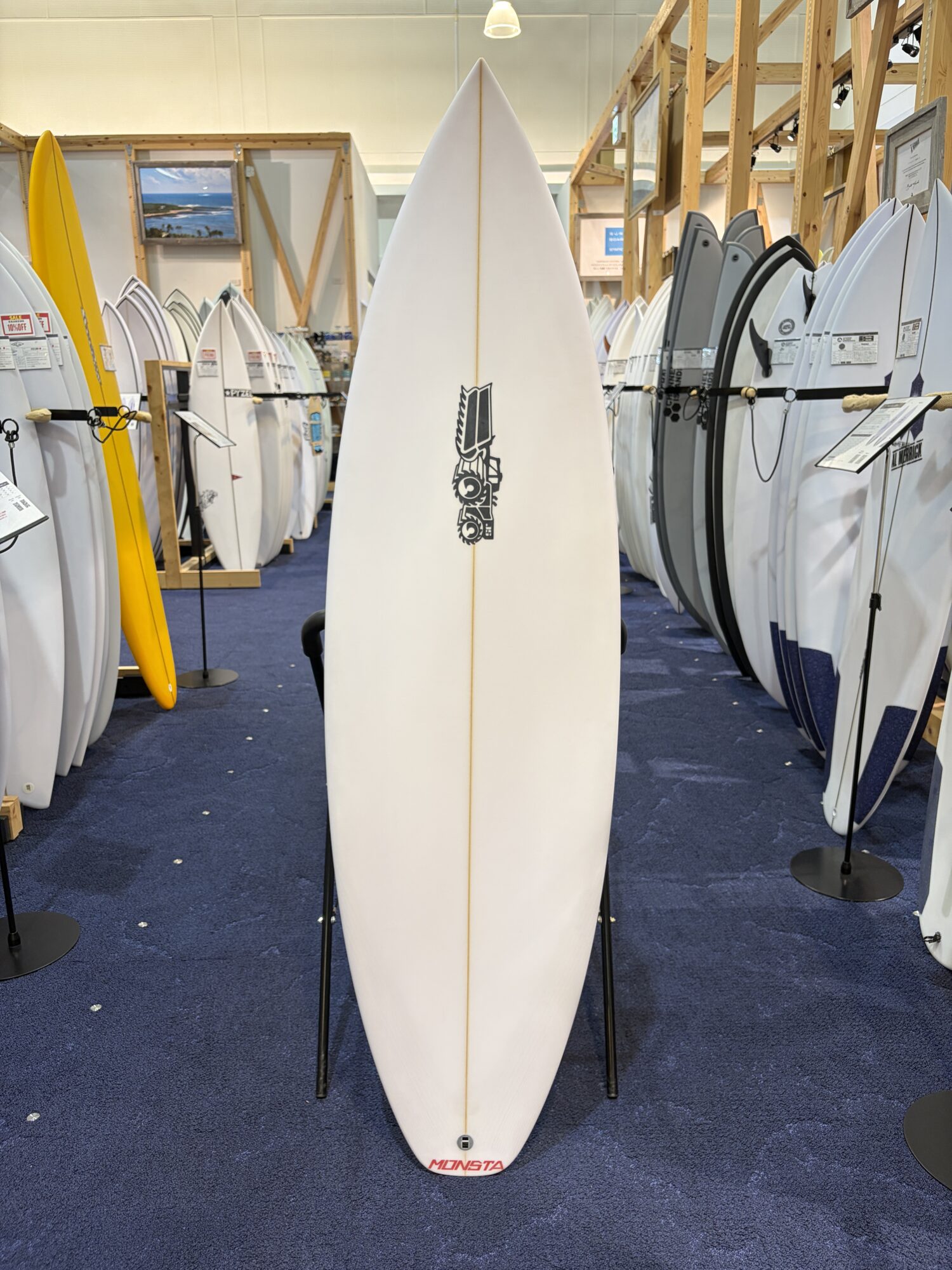 White surfboard with a Monsta logo stands upright on two supports in a showroom filled with surfboards on racks nearby.