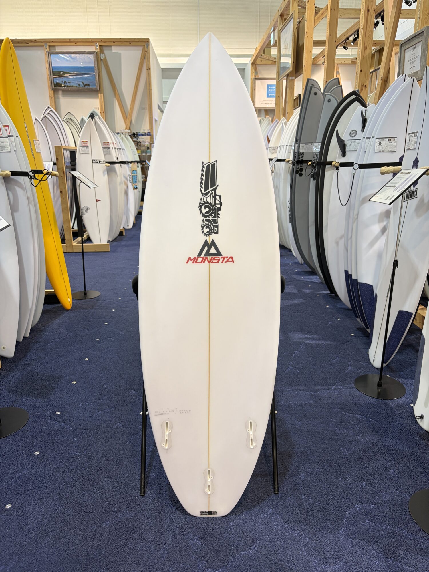 White surfboard on a stand in a showroom, MONSTA logo visible near the bottom center, surrounded by many other boards on racks.