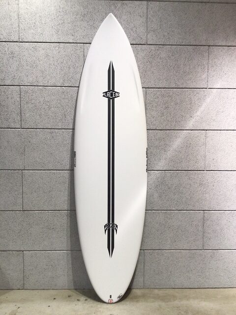 White surfboard with a black vertical stripe and logos, leaning against a gray tiled wall.