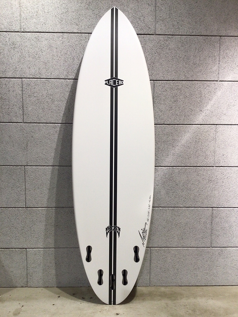White surfboard with black center stripes, a small logo, leaning against a gray brick wall near the floor.