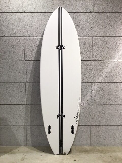 White surfboard with a black center stripe, propped upright against a gray tiled wall; logos along the center and near the tail, fin boxes visible.