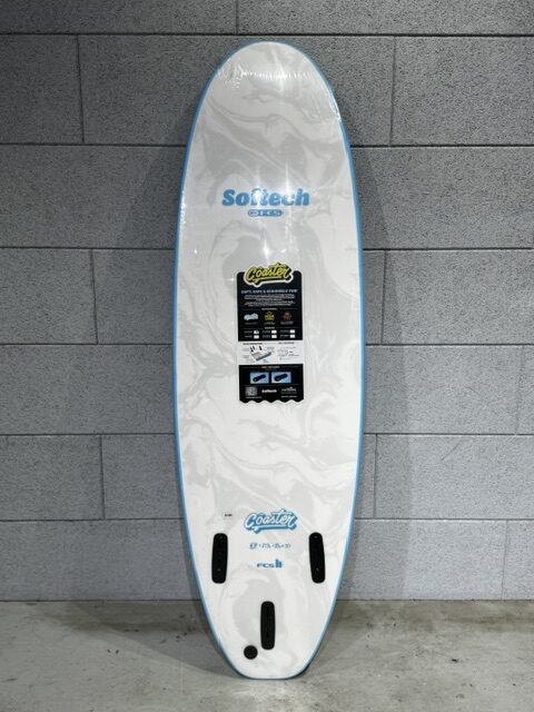 IMG_7065 - SHOP NEWS | ムラサキスポーツオンラインストア 通販 White surfboard with blue edge trim leaning against a gray tiled wall, displaying brand stickers and fin boxes at the bottom.