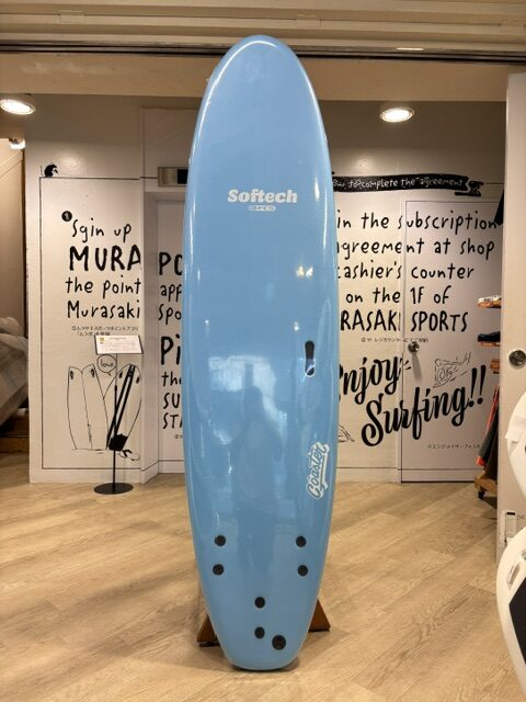 IMG_9653 - SHOP NEWS | ムラサキスポーツオンラインストア 通販 Blue Softtech surfboard on a wooden stand, displayed indoors with shop signage in the background.