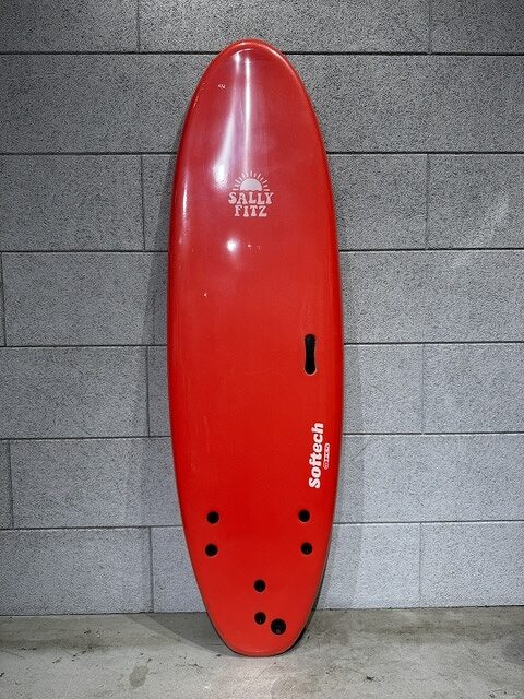s-IMG_3794 - SHOP NEWS | ムラサキスポーツオンラインストア 通販 Red shortboard surfboard leaning upright against a gray brick wall, with visible fin boxes and multiple fin holes near the tail, branded 'SALLY FITZ' and 'Softech'.