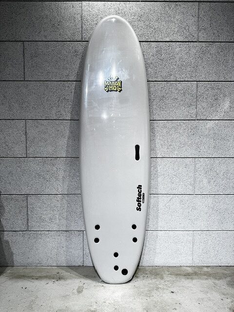 s-IMG_3796 - SHOP NEWS | ムラサキスポーツオンラインストア 通販 White surfboard standing upright against a gray tiled wall, with several tail holes and a fin slot; branding decals visible on the board.