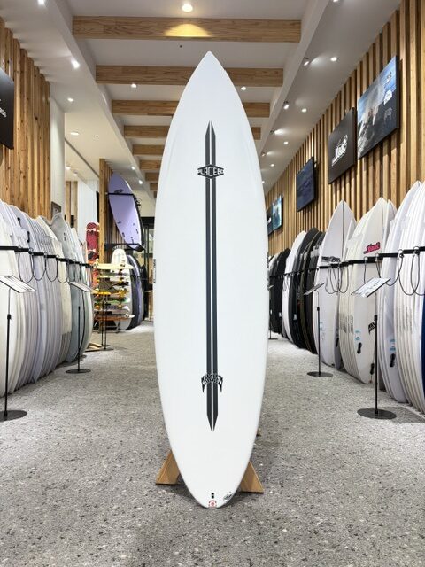IMG_2732 - SHOP NEWS | ムラサキスポーツオンラインストア 通販 Tall white surfboard on a display rack in a surf shop, centered between rows of other boards, with a vertical stripe and logo down the center.