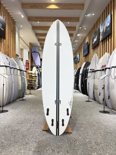 IMG_2733 - SHOP NEWS | ムラサキスポーツオンラインストア 通販 White surfboard with two black stripes standing upright on a wooden rack inside a surf shop showroom, surrounded by rows of other boards on racks.