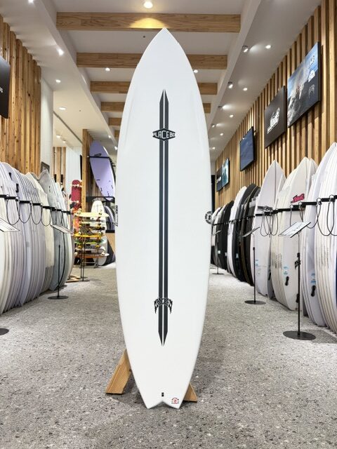 IMG_2734 - SHOP NEWS | ムラサキスポーツオンラインストア 通販 Tall white surfboard with a vertical black stripe and logos stands upright on a wooden rack in a surf shop, surrounded by rows of other surfboards.