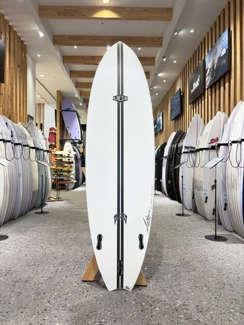 IMG_2735 - SHOP NEWS | ムラサキスポーツオンラインストア 通販 Tall white surfboard with black rails and logo, standing upright on a wooden stand inside a surf shop showroom surrounded by boards on both sides.