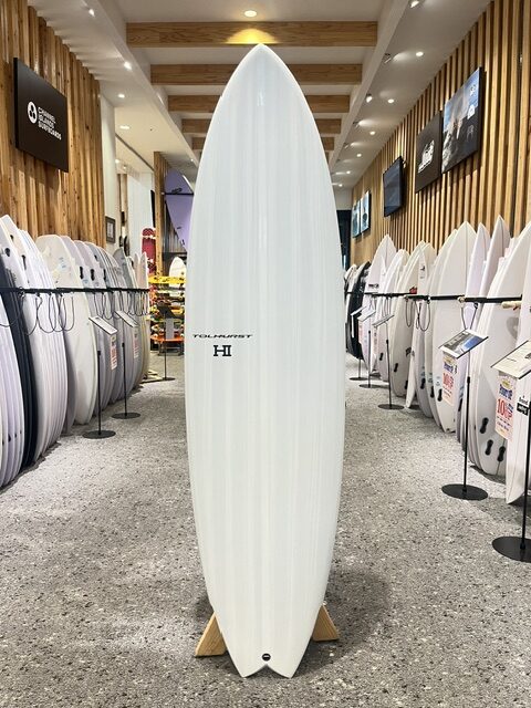 White surfboard standing upright on a display rack in a surf shop, surrounded by rows of other boards on both sides.