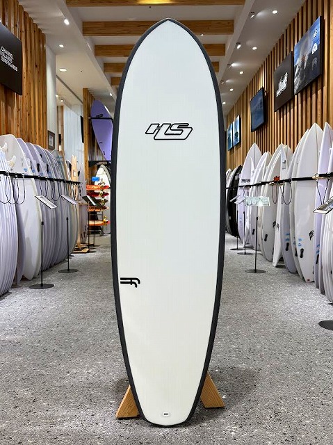 Tall white surfboard with an HS logo stands upright in a surf shop showroom, surrounded by other boards on racks.