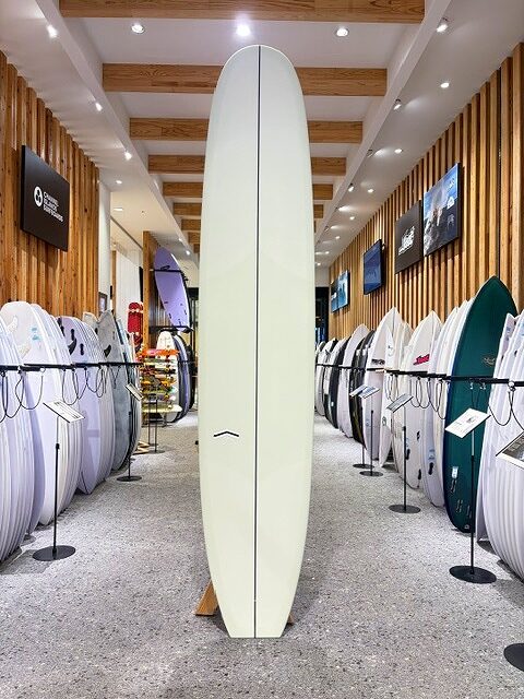 Tall pale yellow surfboard standing upright on a stand in a surf shop, surrounded by rows of other boards on racks with wooden wall panels and ceiling lights.