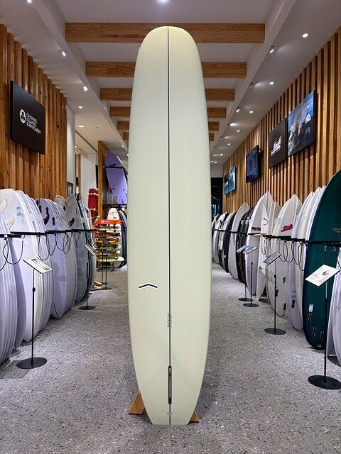 Tall pale-yellow surfboard standing upright on a wooden rack in a surf shop, flanked by rows of boards.