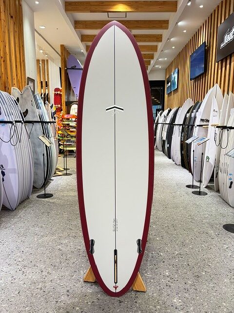 White surfboard with maroon rails displayed upright in a surf shop showroom.