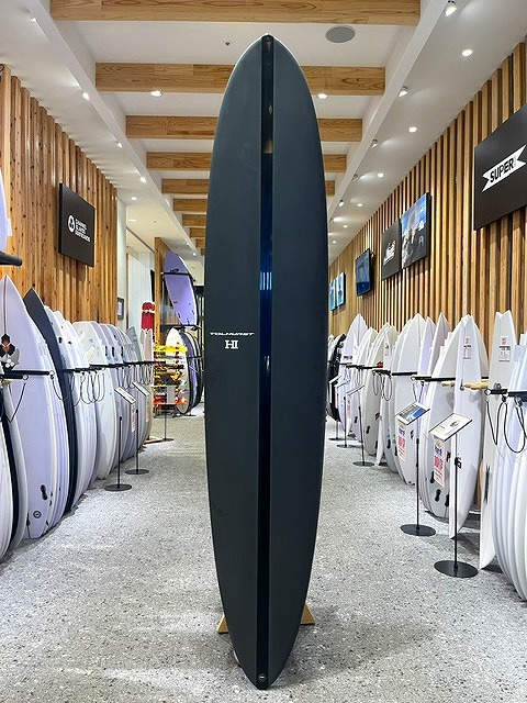 Tall dark blue surfboard standing upright in a surf shop showroom and flanked by other boards.